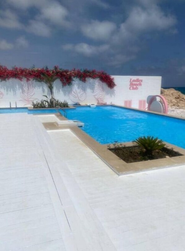 Una piscina con plataforma blanca, rodeada de flores rojas y un letrero que dice 'Ladies Beach Club'.