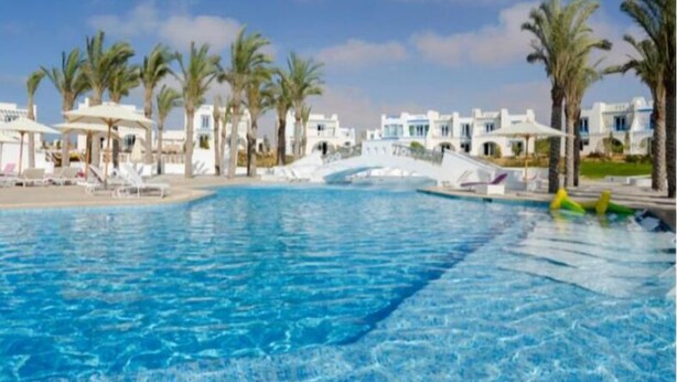 A swimming pool with clear blue water, surrounded by palm trees and white buildings.