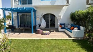 A modern building with a white exterior and blue accents, featuring a covered patio with seating and a hanging chair, surrounded by a well-maintained lawn.