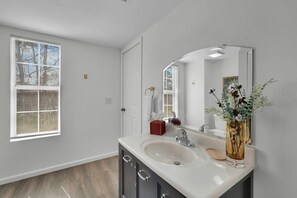 Une salle de bain avec un lavabo blanc, des meubles sombres, un miroir et une fenêtre avec des stores.