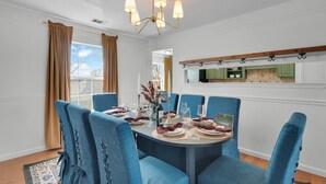 Une salle à manger avec une table ronde dressée pour six personnes, des chaises à revêtement bleu, un lustre et une vue sur la cuisine par une porte ouverte.