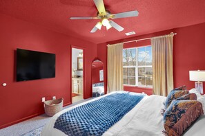 Une chambre avec un mur rouge, un ventilateur de plafond, un lit avec des coussins à motifs, une fenêtre avec des rideaux et un téléviseur fixé au mur.