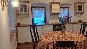Una cocina con zona de comedor, armarios de madera, un ventanal con cortinas y un jarrón con flores sobre la mesa.