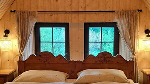 Un dormitorio de madera con techo inclinado, una cama con cabecera de madera, dos ventanas y una lámpara colgante.