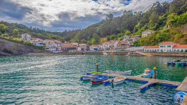 Une ville côtière avec des maisons le long du front de mer, un quai où sont amarrés des bateaux et une colline parsemée de bâtiments.