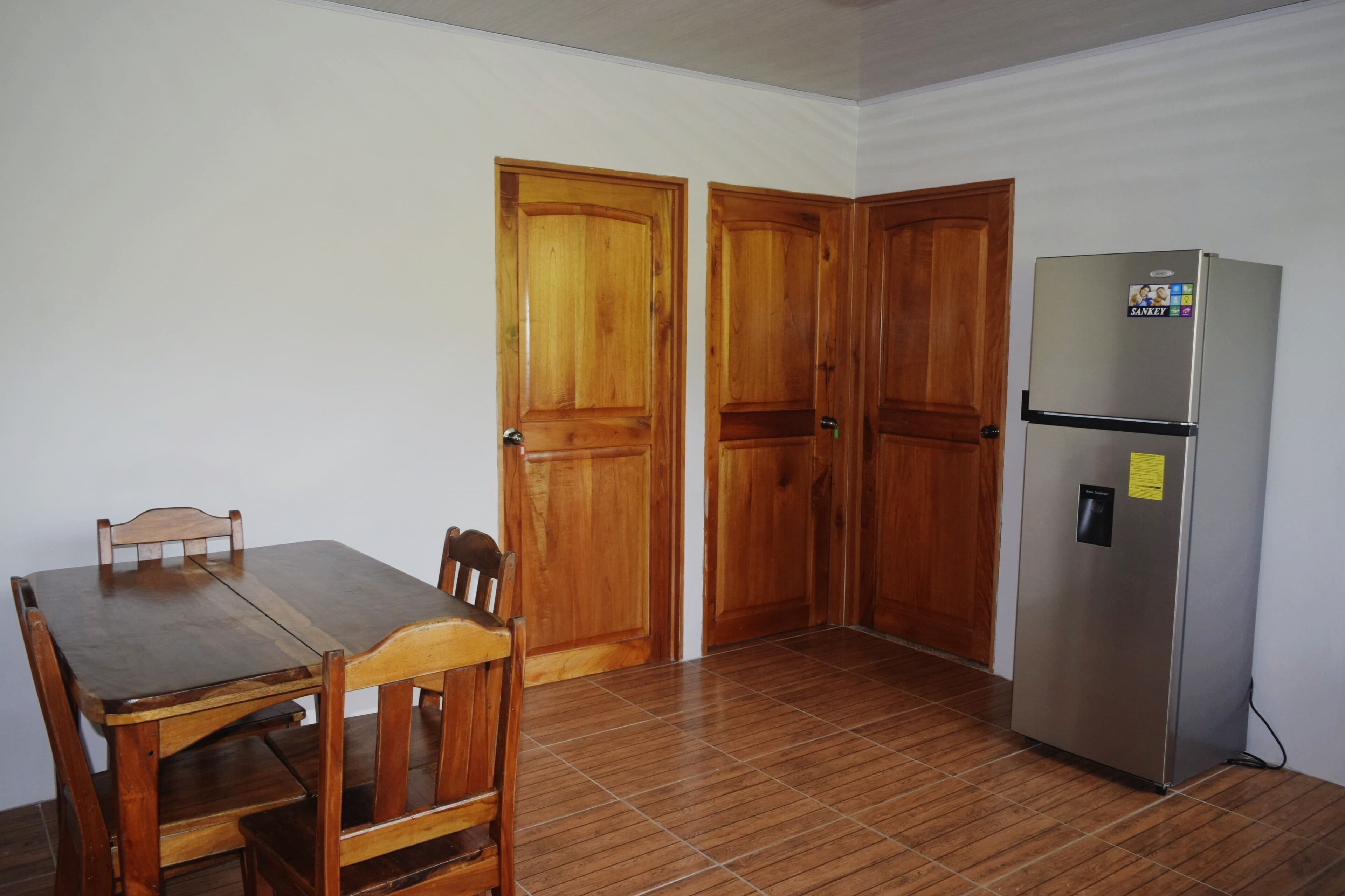 Une pièce comprenant une table et des chaises de salle à manger en bois, deux portes en bois et un réfrigérateur en acier inoxydable.