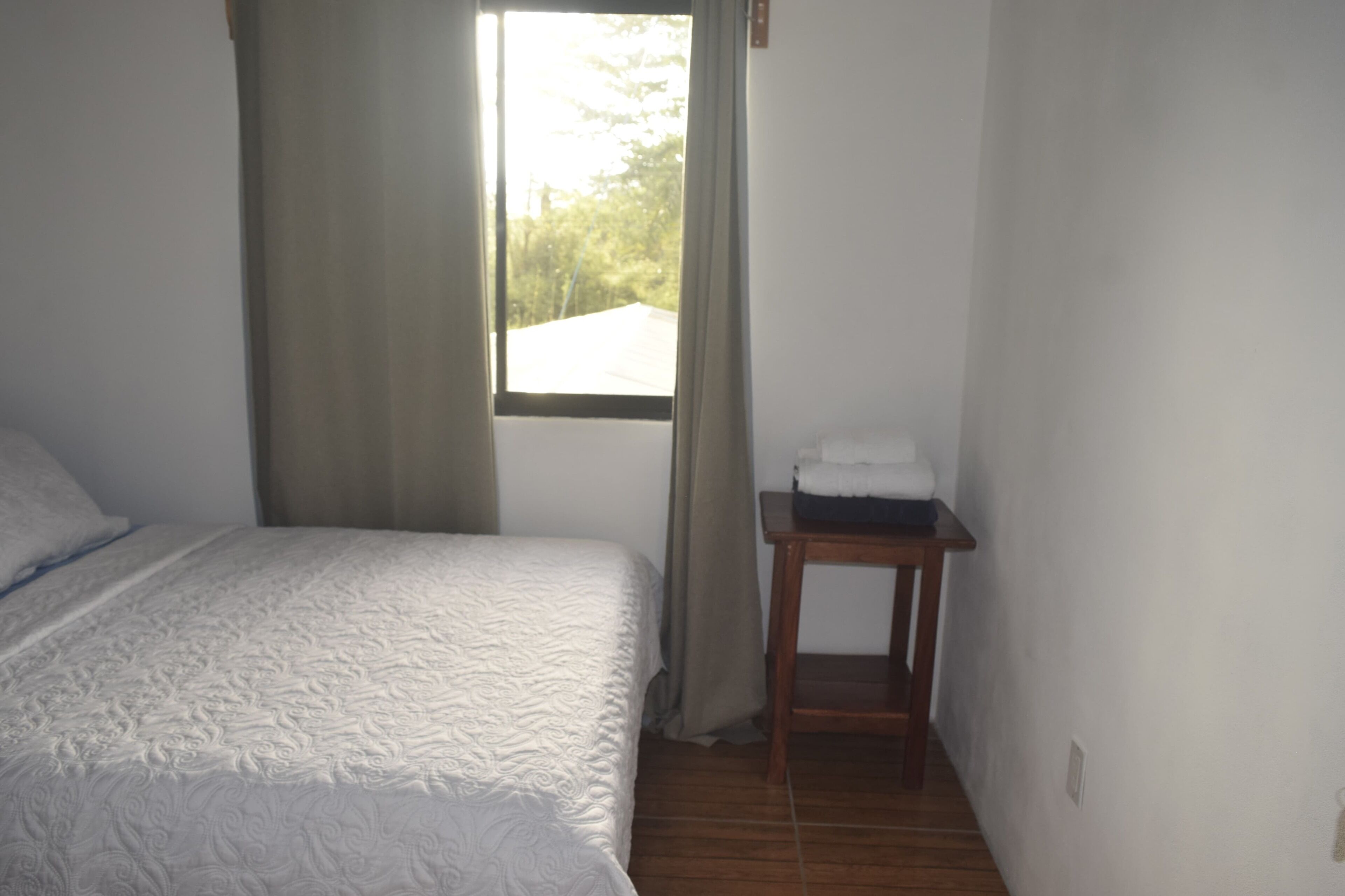 Une chambre avec un lit, une fenêtre avec des rideaux et une petite table en bois sur laquelle est posée une serviette.