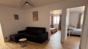 Un salón amplio con un sofá negro, una mesa de centro redonda y piso de madera. Hay un cuadro en la pared y un ventilador de techo.