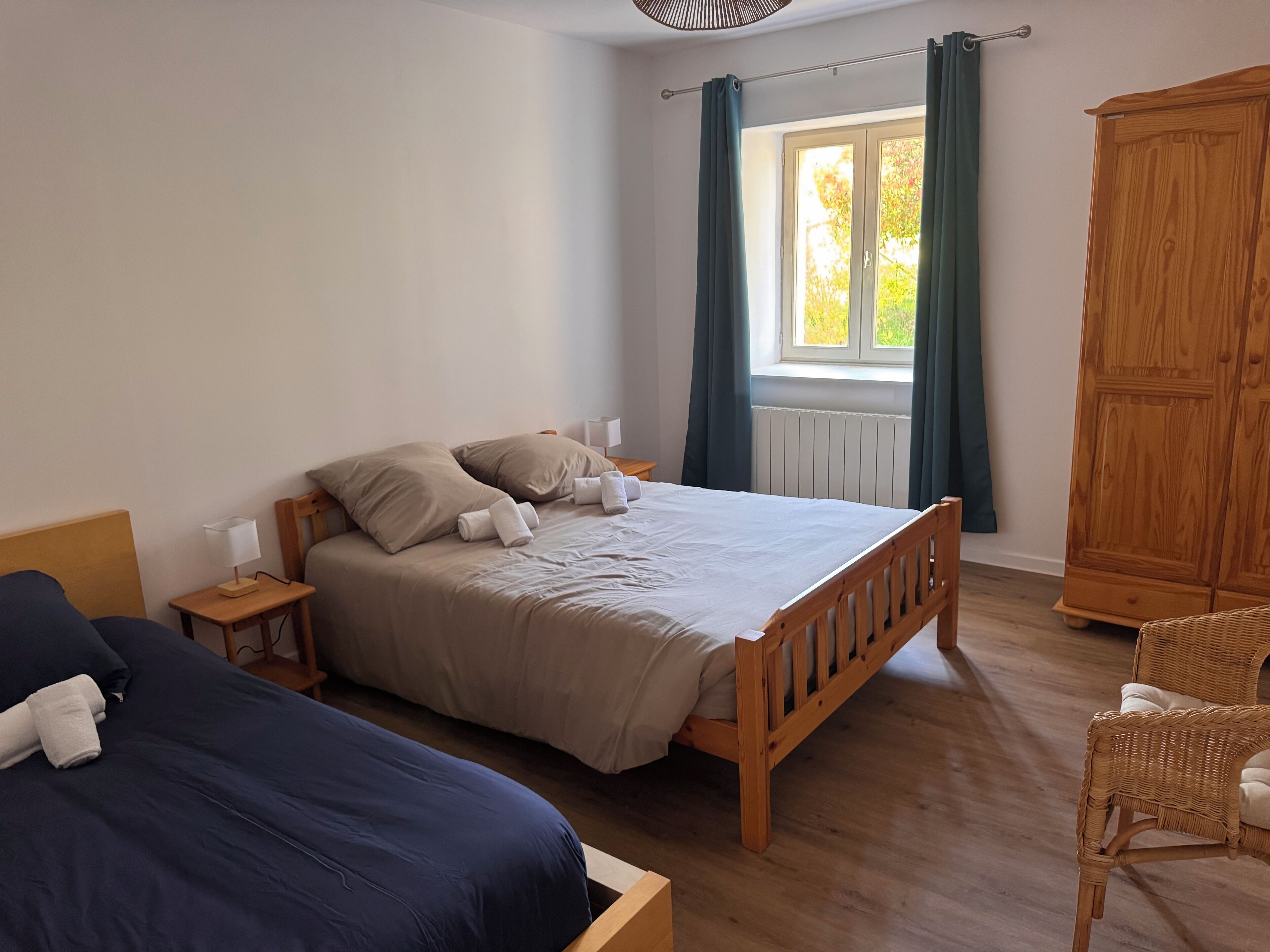 A bedroom with a wooden bed, a nightstand, a lamp, a window with curtains, a wardrobe, and a wicker chair.