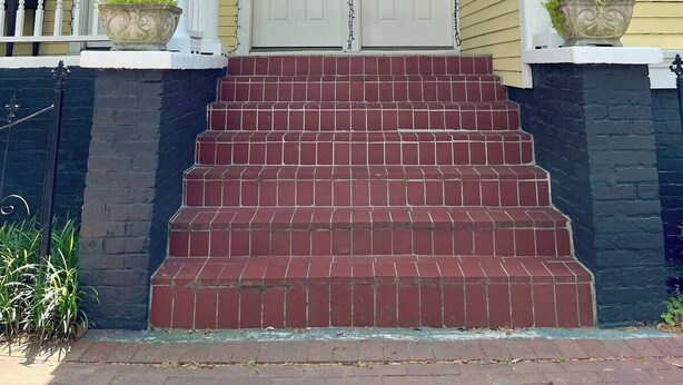 Une maison à deux étages avec un porche à l’avant, une porte blanche et un escalier en briques rouges menant à l’entrée.
