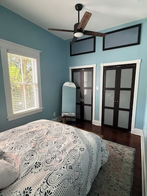 A bedroom with a bed, a ceiling fan, a window with blinds, and a door with frosted glass panels.