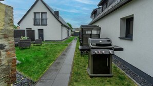 A narrow pathway between two white buildings with a barbecue grill in the foreground.