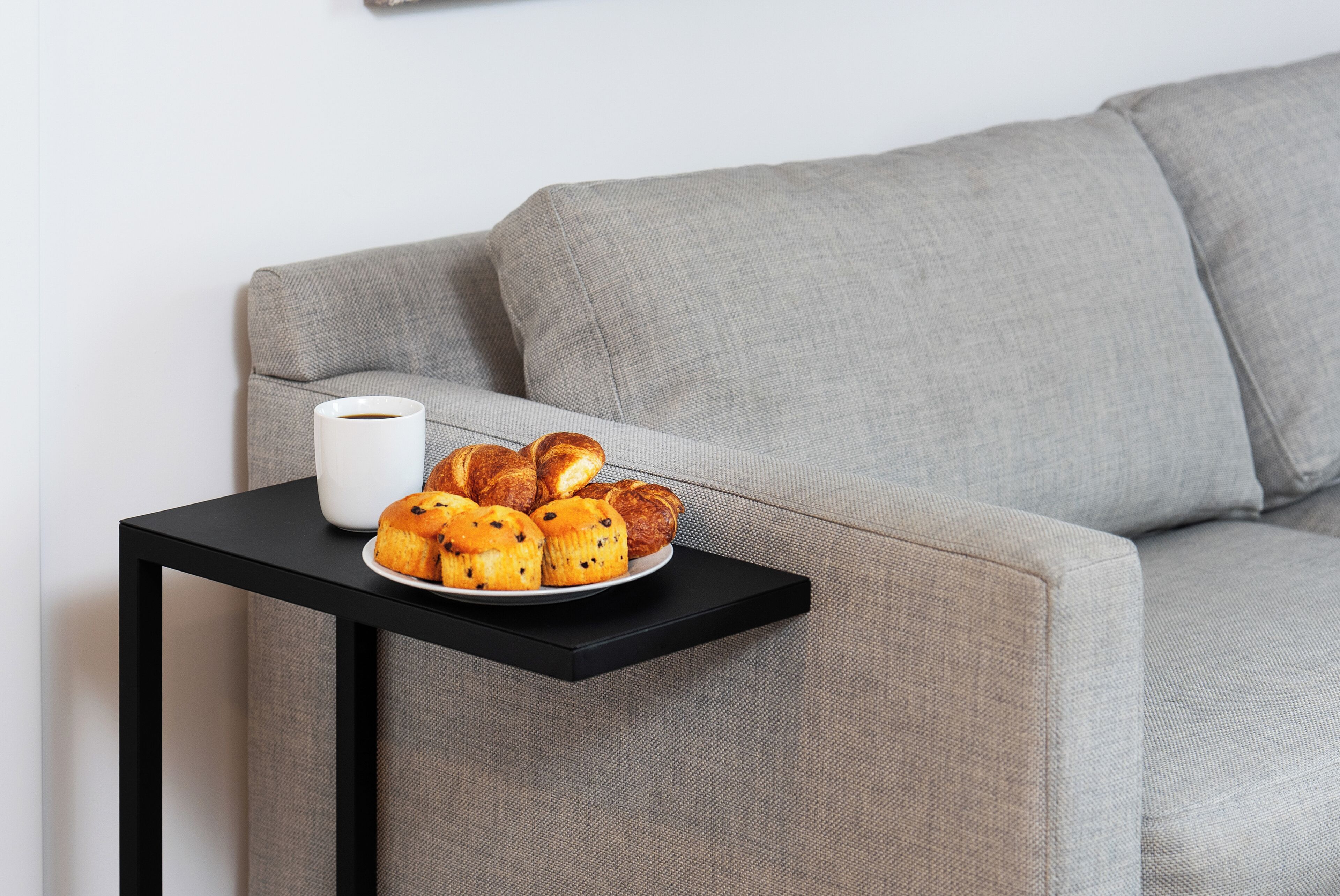 A grey sofa with a black side table holding a plate of muffins and a white mug.