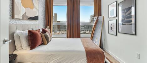 A modern hotel room with a large bed, a view of the city, and artwork on the walls.