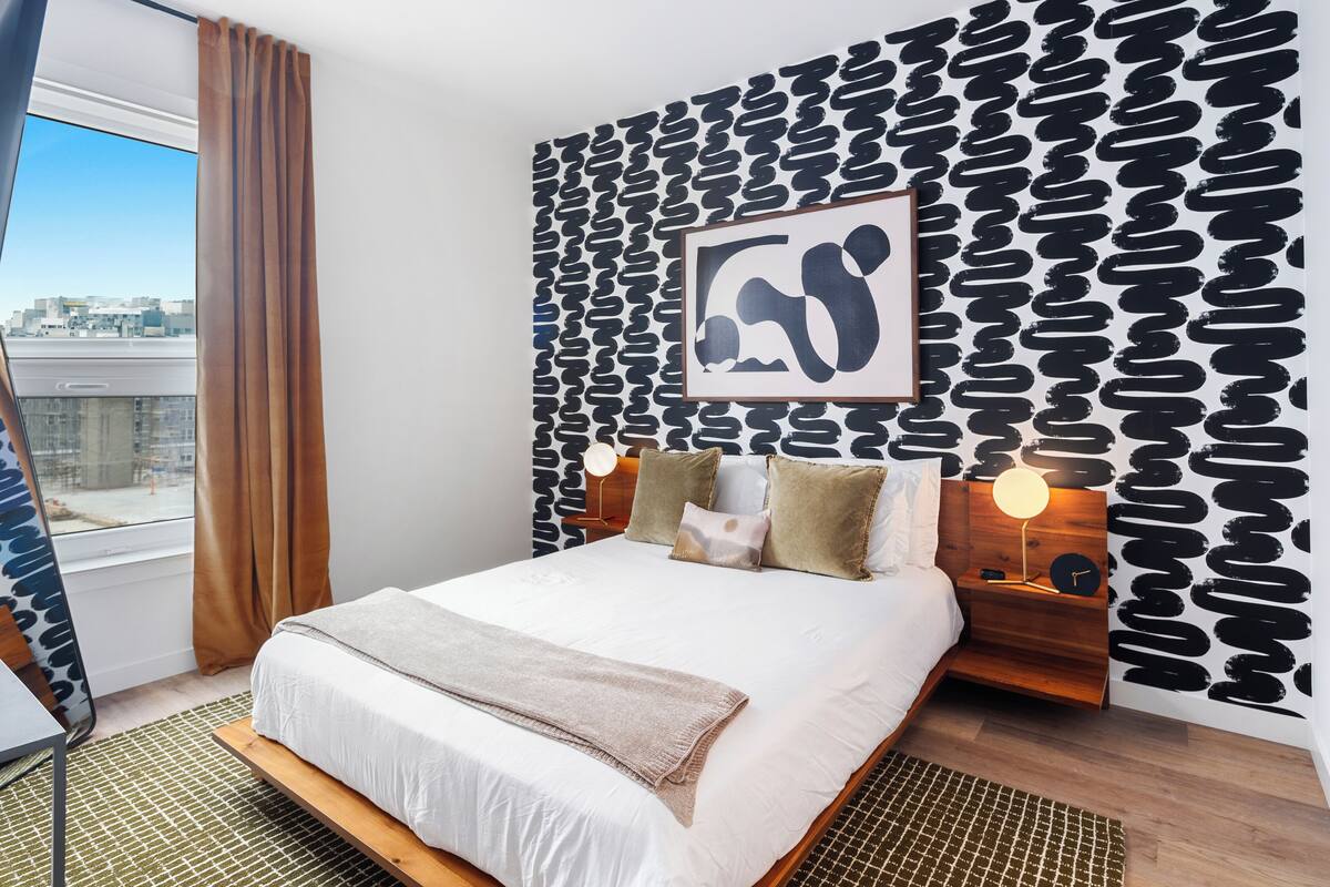 Ein modernes Hotelzimmer mit einem großen Bett, Blick auf die Stadt, einer Wand mit Muster und einem gerahmten Kunstwerk.