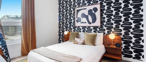 Une chambre d’hôtel moderne avec un grand lit, une vue sur la ville, un mur décoré de motifs et une œuvre d’art encadrée.