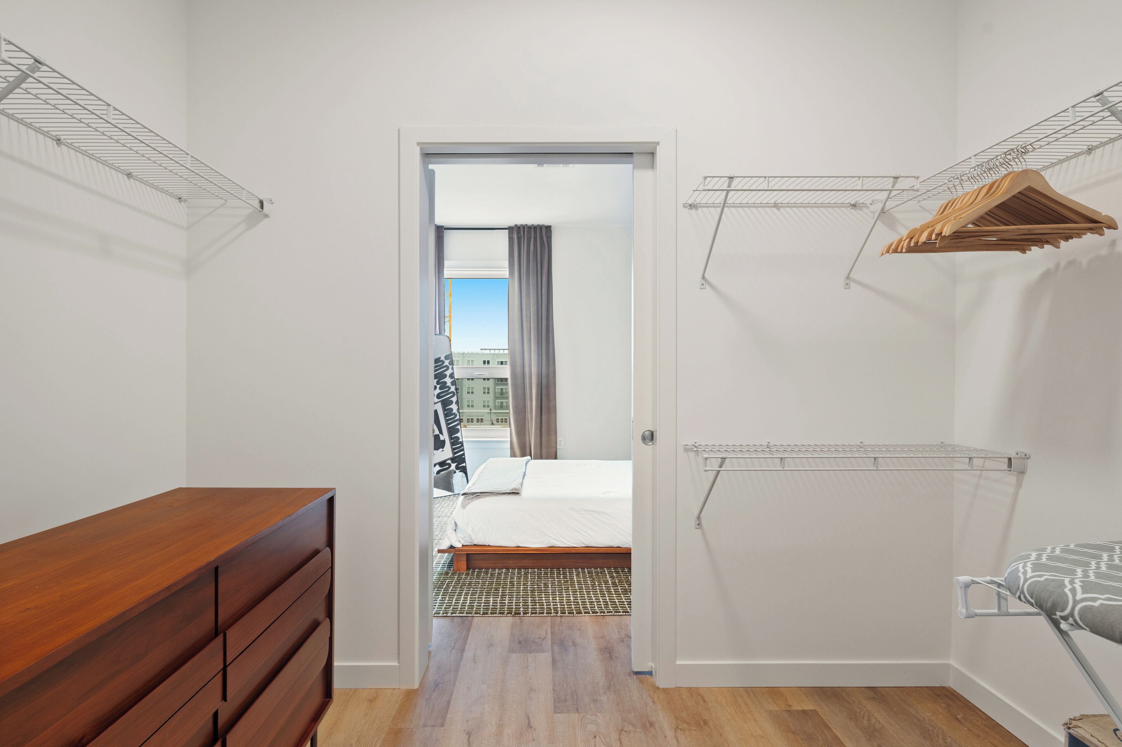 A bedroom with a wooden dresser, a bed, and a view of a cityscape through an open door.