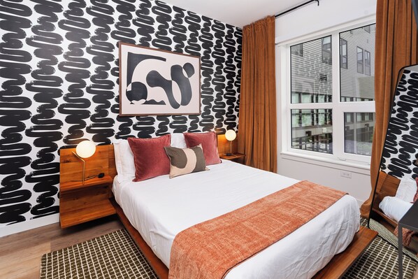 Habitación de hotel con pared estampada, una cama grande con muchas almohadas, una mesita de noche con lámpara y una ventana con cortinas.