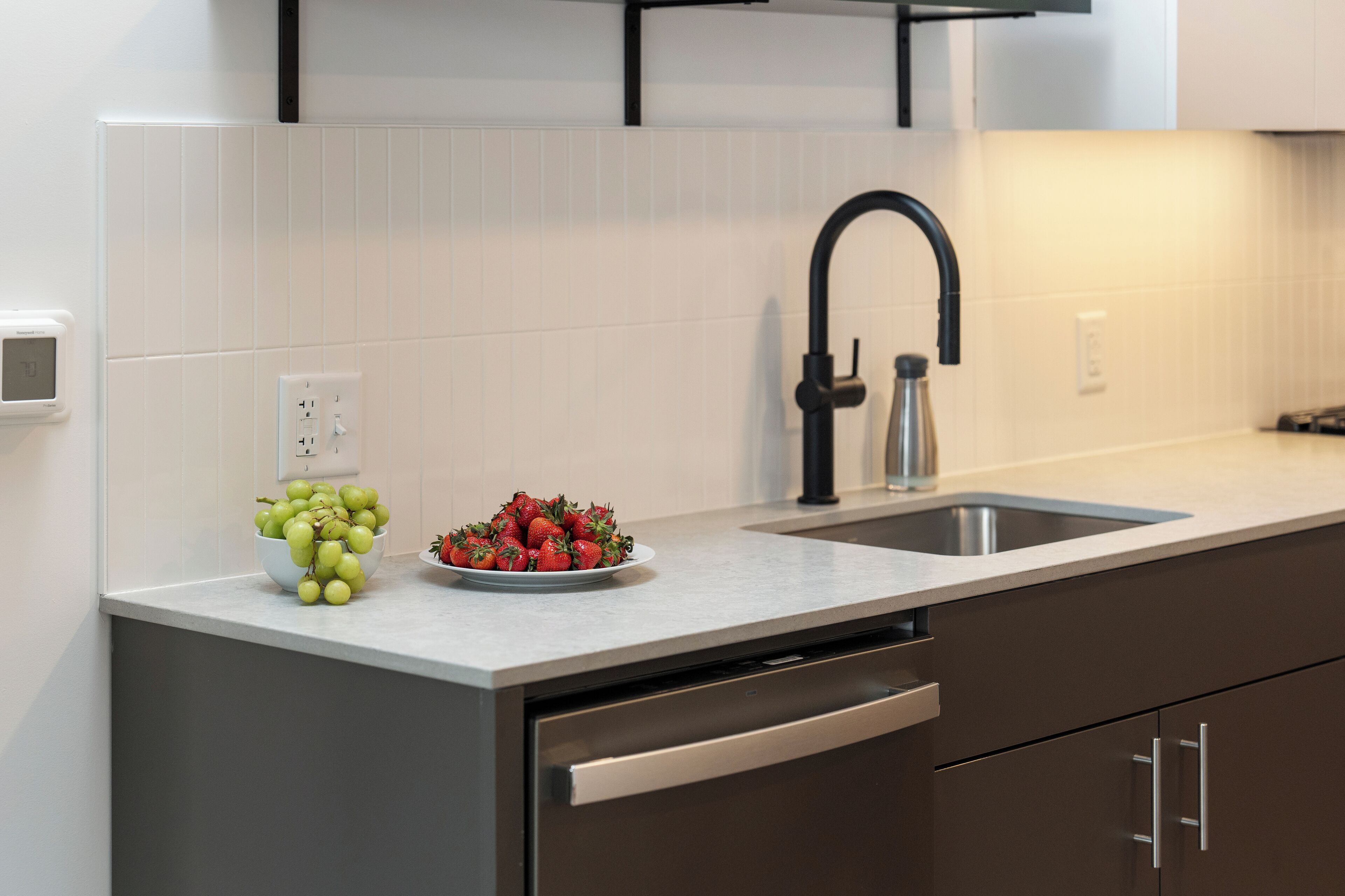 A modern kitchen with a stainless steel sink, a bowl of green grapes, and a plate of strawberries on the counter.