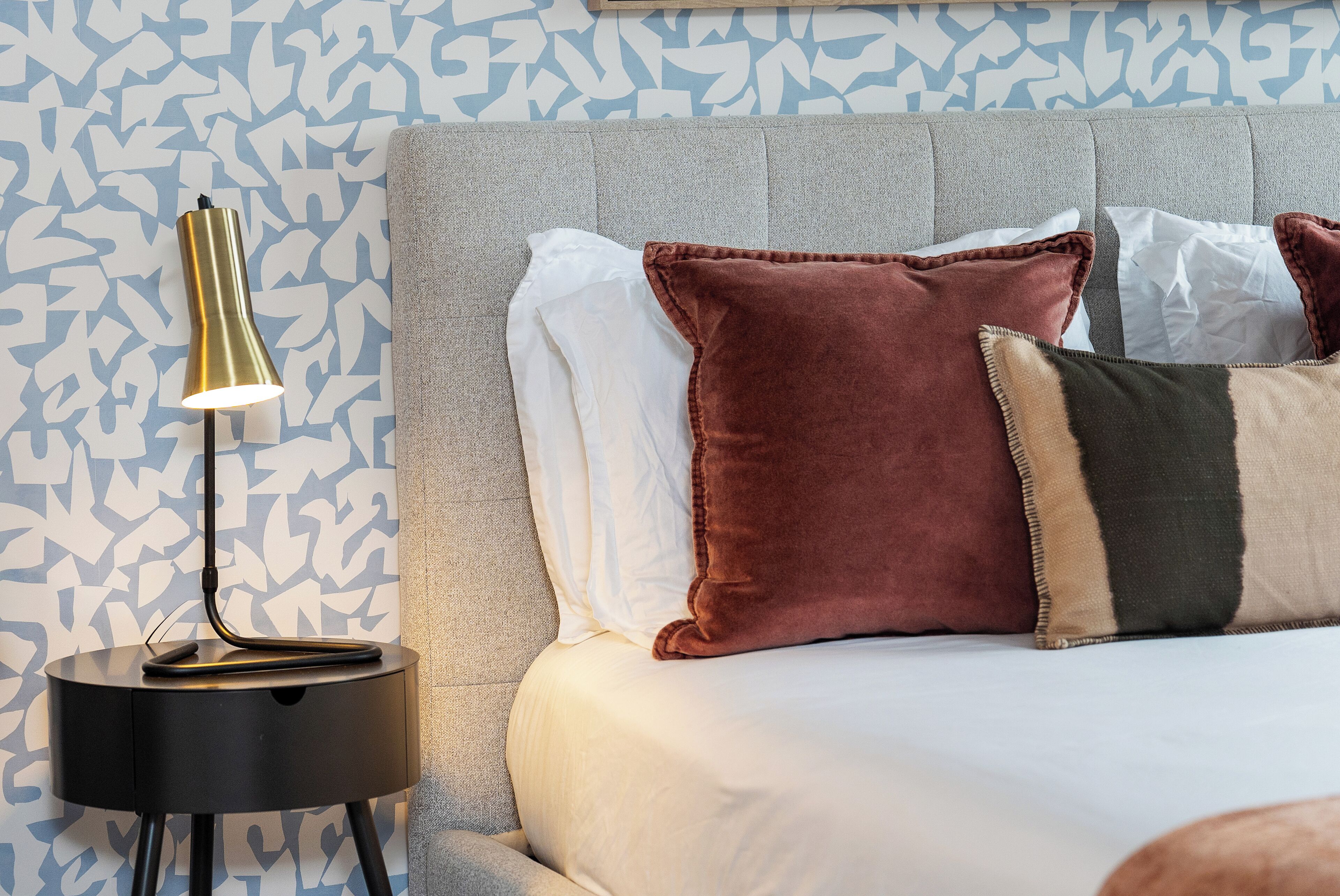 A bed with a grey upholstered headboard, white and brown pillows, and a black side table with a lamp.
