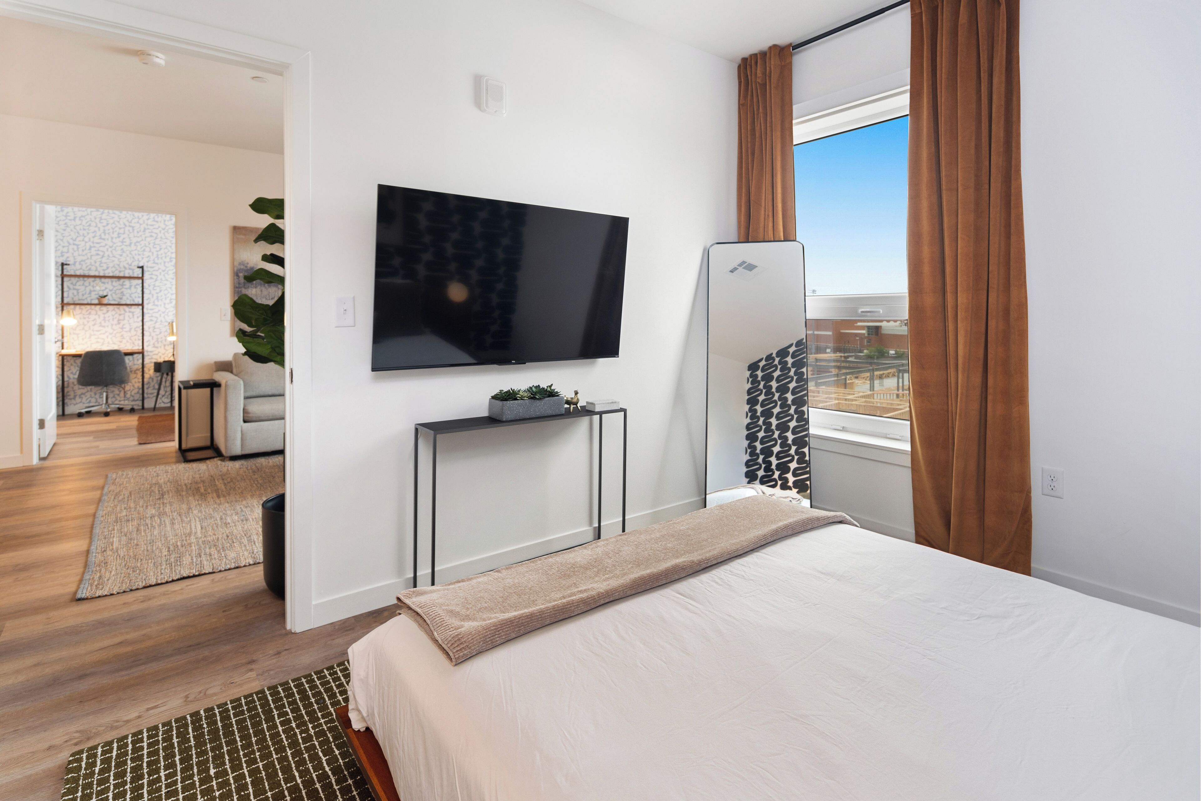 A hotel room with a bed, a TV mounted on the wall, a small table with a plant, and a window with curtains.