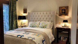 A bedroom with a tufted headboard, floral pillows, a quilt with geometric patterns, a bedside table with a lamp, and a framed picture on the wall. - The Cottage (Mikana)