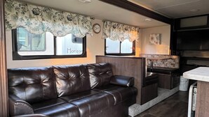 A cozy RV interior with a leather sofa, a small kitchen area, and floral curtains. - Wildwood glamping rv - Site #72 (Klamath)