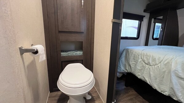 A compact bathroom with a toilet, a small shelf, and a bed with a white bedspread.