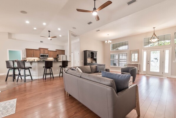 A spacious living area with a large grey sectional sofa, a kitchen with wooden cabinets, and a dining area with bar stools. - Wow! Ultra-spacious 4/2 pet-friendly beach home w/pool, fenced-in yard, sleeps 8 (New Smyrna Beach)