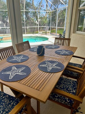 A wooden table with star-shaped placemats, surrounded by chairs with patterned cushions, set in a covered outdoor area with a view of a swimming pool. - Wow! Ultra-spacious 4/2 pet-friendly beach home w/pool, fenced-in yard, sleeps 8 (New Smyrna Beach)