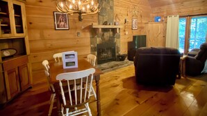 Een gezellig interieur van een chalet, met een houten eettafel, witte stoelen, een open haard en een kroonluchter.