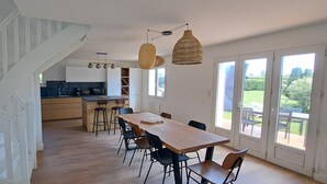 A spacious dining area with a long wooden table and chairs, a kitchen in the background, and a staircase leading to another level. - Renovated, detached belle-illoise family home, 3 bedrooms, sleeps 9 (Le Palais)
