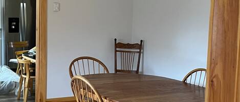 Een eetgedeelte met een houten tafel en vier stoelen, een houten vloer, en een keuken met een roestvrijstalen koelkast.