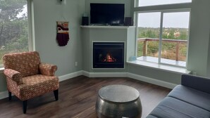 Una sala de estar con chimenea, un sofá, un sillón estampado, un televisor sobre una estantería y un balcón con vistas a zonas verdes.