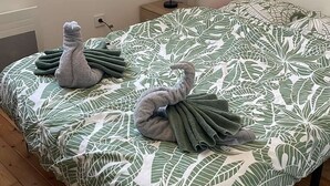 Ein Schlafzimmer mit einem Bett, das mit einer bettwanddecke mit Blättermotiv bezogen ist, einem Paar gefalteter Handtücher auf dem Bett, einem Nachttisch mit einer Lampe und einem Fenster, das natürliches Licht hereinlässt.