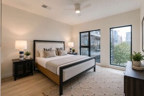 A modern bedroom with a large bed, two bedside tables with lamps, a view of the city through large windows, and a ceiling fan.