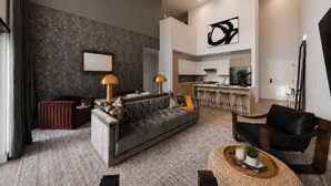 A modern living room with a grey sofa, a coffee table, and a kitchen area with a bar. - Loftline Social - 8BR | 6BA | Sleeps 20 (Nashville)