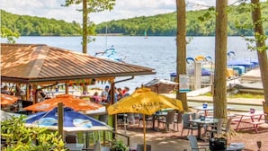 An outdoor seating area with tables and umbrellas, overlooking a lake with boats and a forested shoreline. - Big Boulder Relaxation, Slopes Up, Big Boulder, Jack Frost Ski Resorts  (Lake Harmony)
