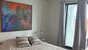Un dormitorio con cama, ventilador de techo, un cuadro abstracto grande y vistas a través de un ventanal.