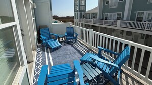 Un balcón con sillas Adirondack azules y una mesita, con vistas a una zona residencial con una cerca blanca y un edificio verde.
