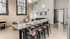 A modern dining area with a long table set for six, surrounded by bar stools. There are large windows with blinds, a ceiling with exposed ductwork, and a refrigerator to the right.