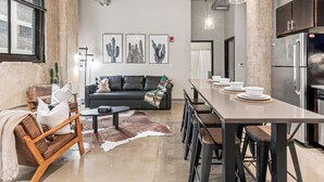 A modern kitchen with a long countertop and bar stools, a black sofa with decorative pillows, and a dining table with white plates.