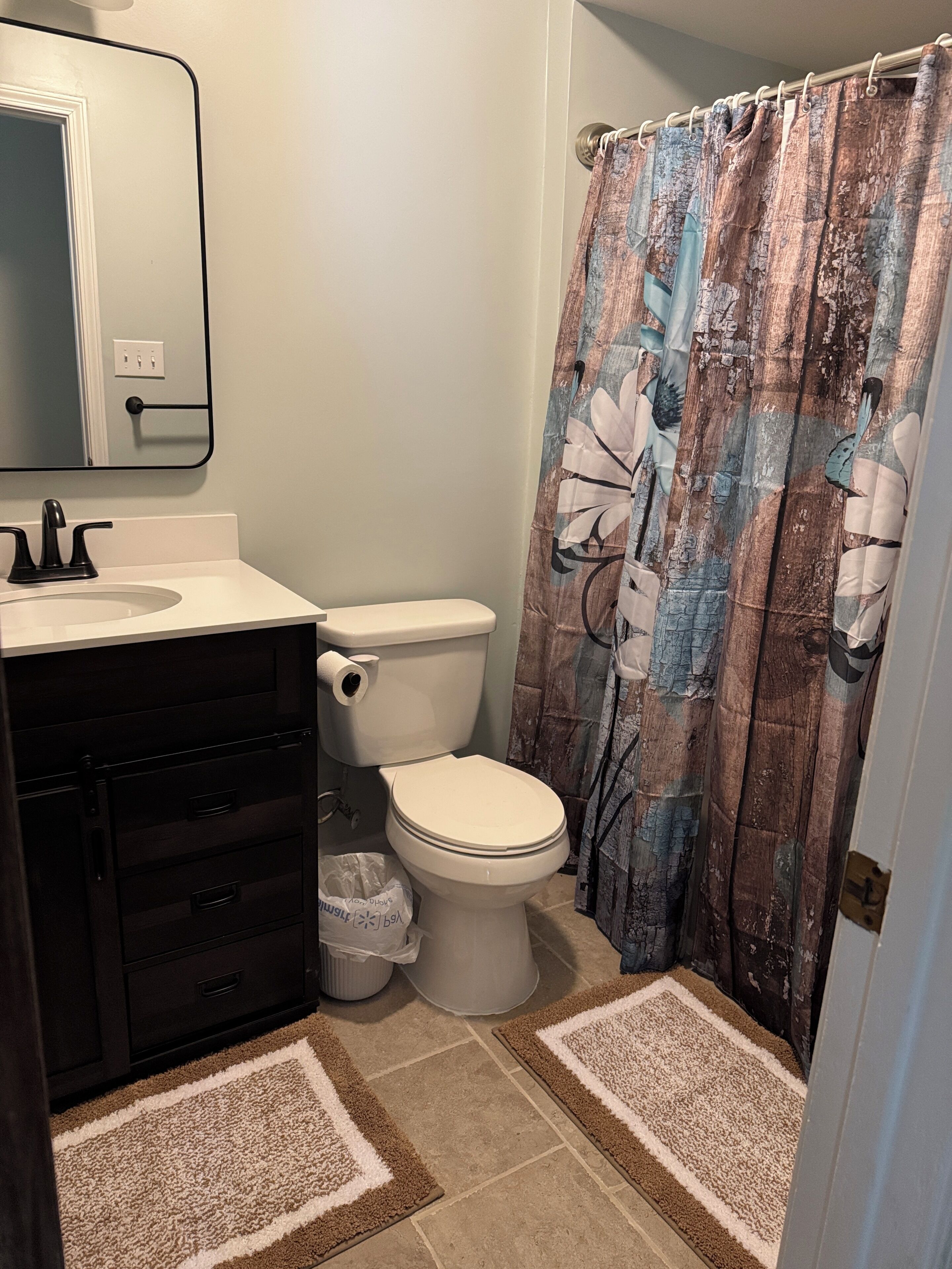 Un baño con inodoro blanco, tocador negro con cajones, espejo y cortina de ducha con motivos florales.