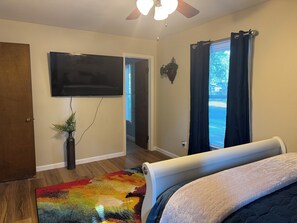 Een slaapkamer met een bed, een aan de muur bevestigde televisie, een plafondventilator en een raam met gordijnen.