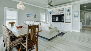 A modern living room with a dining area, white furniture, a chandelier, ceiling fan, and built-in entertainment unit. - The Lazy Turtle, Beachfront (Crystal Beach)