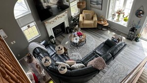 Ein modernes Wohnzimmer mit einer Eckcouch, einem Kamin, einem Fernseher und großen Fenstern.