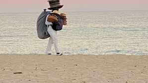 Una persona de pie en la playa con un montón de sombreros y una mochila, mirando hacia el océano.
