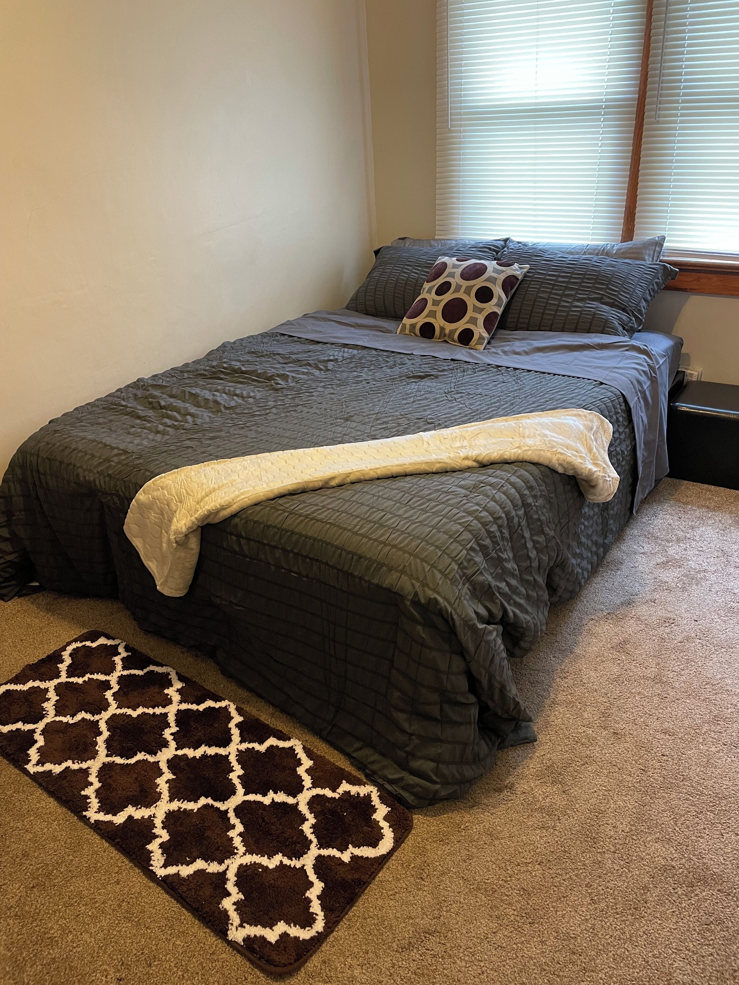 A bedroom with a bed, a window with blinds, a carpet, and a rug.