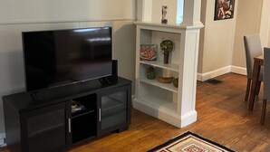 A living room with a flat-screen TV, a black entertainment unit, a white shelving unit with decorative items, a patterned rug, and a wooden coffee table with a remote control. - Charming 2-bedroom house in busy downtown Indianapolis with WiFi, AC. (Indianapolis)