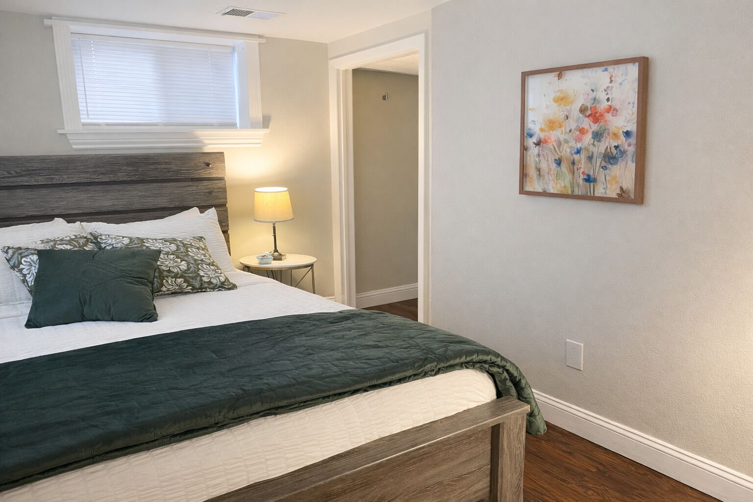 Un dormitorio con cama, mesita de noche, lámpara, ventana y un cuadro en la pared.